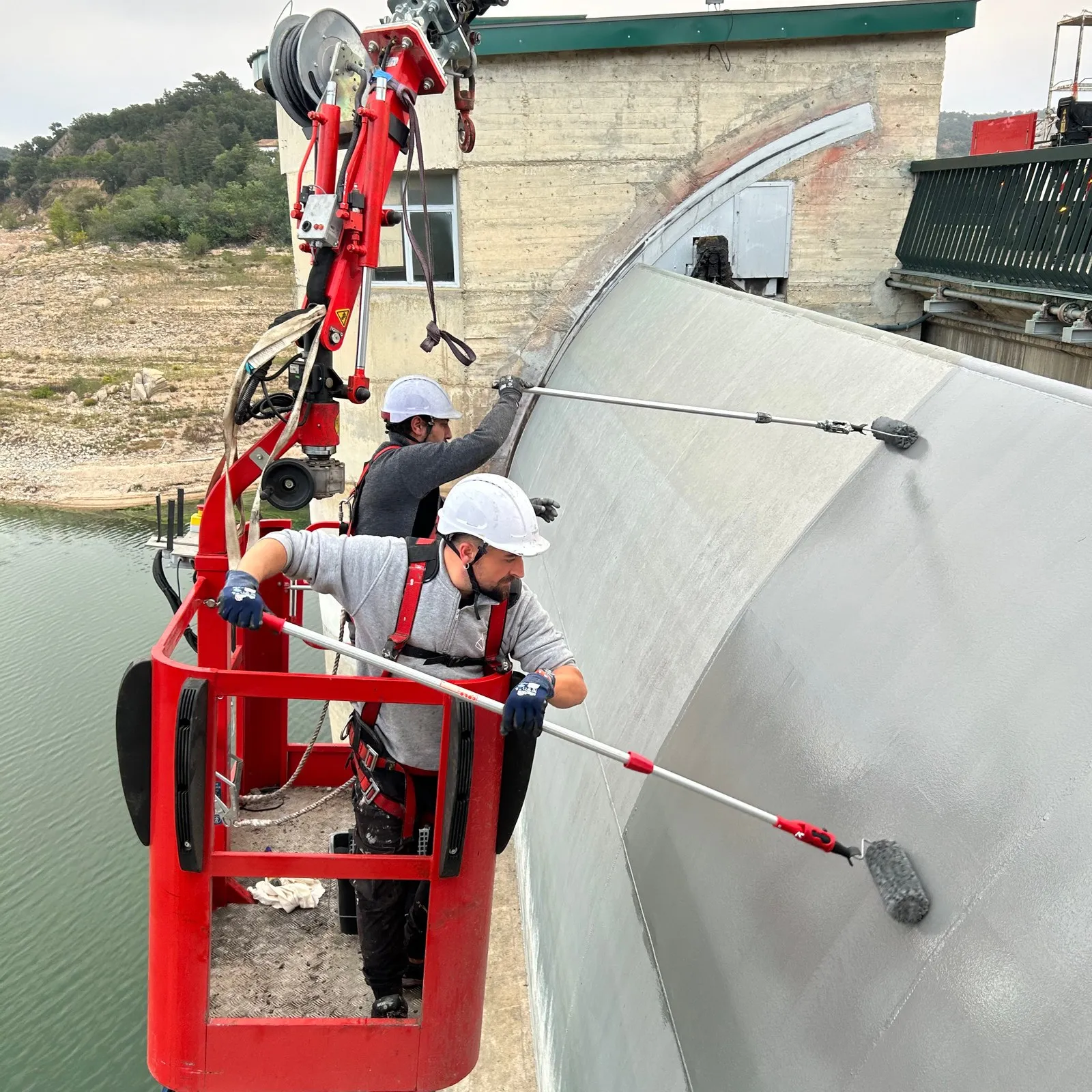 Operaris pintant el mur de formigó d'una presa des d'una cistella elevadora